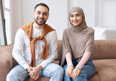 Evdeki oturma odasındaki kameraya gülümseyen neşeli Müslüman çift. İki kişilik modern Arap Ailesi 'nin portresi, Mutlu Genç karı-koca başörtüsü içinde birlikte poz veriyorlar.