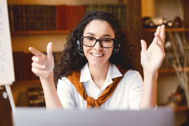 Sanal Öğretmenlik. Gözlük takan, kablosuz kulaklık takan, video konuşması yapan, yeni tema ya da konu öğreten evde çevrimiçi ders sırasında kamerayla konuşan pozitif genç bir bayan profesör.