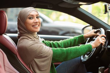 Şehir turu. Türbanlı Mutlu Müslüman Kadın Modern Araba Kullanıyor, Direksiyon Tutuyor 'un yan görünümü. Orta Doğulu kadın yeni otomobil için test sürüşü yapıyor, kameraya bakıyor ve poz veriyor.