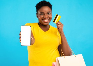 Mobil Alışveriş. Gülümseyen Afrikalı Kadın, telefon ekranı ve kredi kartı pozu gösteriyor. Mavi Stüdyo arka planında alışveriş torbası var. Kadın Alıcı Akıllı Telefon Bankacılık Uygulamasını Öneriyor. Model
