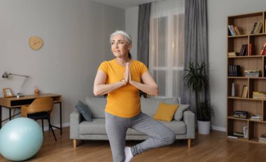 Yurt içi yoga pratiği. Yaşlı kadın ağaç duruşu yapıyor, dengesini koruyor, evde oturma odasında antrenman yapıyor. Spor giyim sektöründe spor salonunda egzersiz yapan mutlu bir kadın.
