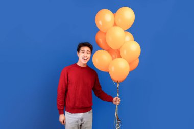 Tatil kavramını kutluyoruz. Elinde sarı balonlar tutan heyecanlı Asyalı genç adamın dikey çekimleri. Mavi arka plan ve afiş üzerinde izole edilmiş pozlar. Gülümseyen adam partiye hazır.