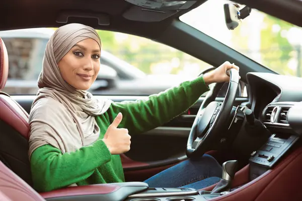 Ortadoğu 'dan kendine güveni tam genç bir kadının portresi. Başörtüsü takıyor. Gösterişli arabanın içinde oturan el hareketlerini gösteriyor. Gülümseyen Müslüman Kadın Müşteri İyi İşaret Veriyor, Ehliyetini Aldı
