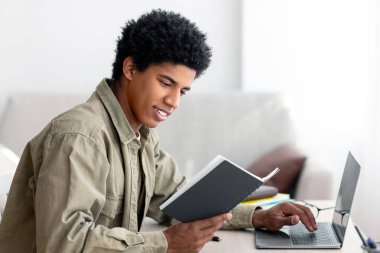 E-öğrenme konsepti. Yakışıklı siyahi genç notlarını ev ödevini evde bilgisayarla yapmak için kullanıyor. Pozitif Afro-Amerikan ergen uzaktan eğitim alıyor, internet dersi alıyor.