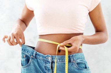 Siyah, büyük kot pantolonlu, belini bantla ölçen, zayıflatma diyeti veya yağ aldırma sonucu ortaya çıkan, gri arka plan. Genç bayan kilo vermek için sağlıklı beslenmeyi teşvik ediyor.