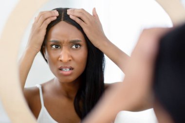 İlk gri saçlar, yaşla ilgili değişiklikler ve evde bakım. Milenyum hüzünlü afro-amerikalı kadın pürüzsüz saçlara dokunuyor ve aynaya bakıyor sabah yatak odasının içinde, fotokopi odasında, kapalı alanda