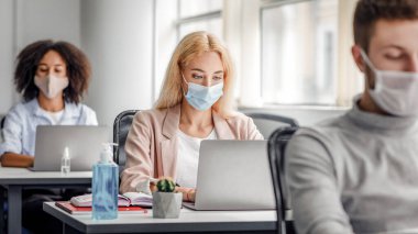 Karantina salgını sırasında iş yerinde sosyal mesafe ve yeni normallik. Koruyucu maskeli çok ırklı adam ve kadınlar antiseptikli masalara dizüstü bilgisayarlara bakarlar.