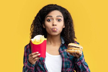 Fast Food aşığı. Burger yiyen, patates kızartması yiyen, ağzında cips tutan komik Afrikalı Amerikalı kadın portresi. Siyah Bayan Sandviç ve Lezzetli Patates, portakal arkaplanda izole edilmiş, bayrak.