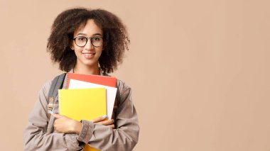 Modern öğrenci, karantina sonrası zeki ve inek öğrenci. Sırt çantalı, gözlüklü, gülümseyen Afrikalı Amerikalı genç kadın not defterlerine sarılıyor ve sarı arka planda izole edilmiş kameraya bakıyor.