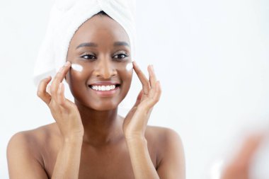 Güzellik blogcusu, nemlendirici ve yaşlanmayı önleyici cilt bakımı. Milenyum güzeli, neşeli Afro-Amerikan kadın sabah evde yüzüne krem sürüyor ve yatak odasındaki aynaya bakıyor.