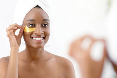 Modern kozmetik ürünleri, evde günlük kaplıca ve yaşlanma karşıtı bakım. Duş aldıktan sonra beyaz havluyla, gözlerinin altına altın bantlar takıp yatak odasının iç tarafındaki aynaya bakan neşeli, Afro-Amerikan kadın.