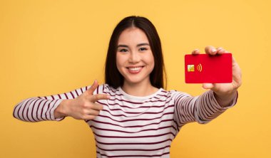 Uzun siyah saçlı neşeli bir Asyalı kadın onay verirken temassız bir ödeme kartı gösteriyor. Parlak sarı arka plan onun ifadesine canlı bir atmosfer ekler..