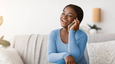 Evde cep telefonuyla konuşan Afro-Amerikalı olumlu bir kadın arkadaşlarıyla ya da aile telefonuyla rahat bir oturma odasında dinlenirken sohbet ediyor.