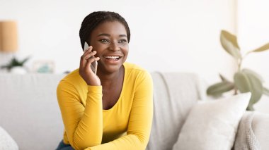 Telefon var. Neşeli genç Afrikalı Amerikalı kadın evde cep telefonuyla konuşuyor, arkadaşlarıyla konuşuyor ve gülümsüyor, keyifli sohbetler yapıyor, mobil iletişimin tadını çıkarıyor, koltukta oturuyor, boş bir alanda oturuyor.