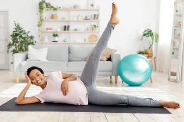 Müstakbel zenci kadının yoga minderinde egzersiz yaparken, bacağını kaldırırken, evde pozitif ve sağlıklı hissederken boy boy boy portresi. Hamile Afrikalı Amerikalı kadın oturma odasında çalışıyor.