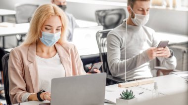 Ofisteki müşterilerle çalışmak için modern aletler. Milenyum maskeli bir adam akıllı telefondan yazı yazıyor. Kadın iş yerinde bilgisayarına bakıyor. Cavid-19 salgını sırasında koruyucu camla.