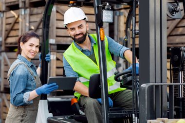 Depo işçileri, modern teknolojiyle endüstri yönetimi. Tabletli gülümseyen milenyum kadını, forklift siparişi veren kasklı sakallı bir adamla konuşuyor.