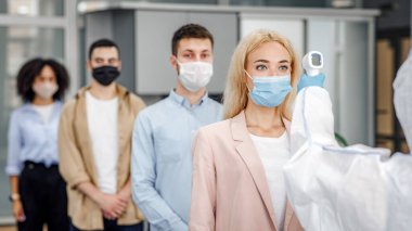 İş yerinde sıcaklık kontrol testi ve sosyal uzaklık. Koruyucu giysili adam, sabahleyin ofise girerken çok uluslu çalışanları temas olmayan termometreyle kontrol ediyor.