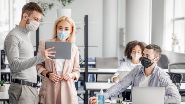 Şirket şirketinde takım çalışması ve koronavirüs salgını sırasında sosyal mesafe. Koruyucu maskeli erkek yönetici, ofis içi çalışanlarına tablet, dizüstü bilgisayarlar ve masalarda antiseptik gösteriyor.