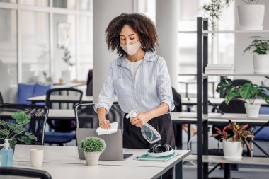 Koronavirüsü önleme. Koruyucu maskeli Afrikalı Amerikalı kız bilgisayarını sünger ve spreyle dezenfekte ediyor.