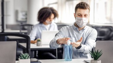 Coronavirüs salgını sırasında güvenlik kuralları. Koruyucu maskeli bir adam iş yerinde ellerini antiseptikle tedavi ediyor. Modern çalışma odasının içinde bilgisayarla, kopyalama alanında.