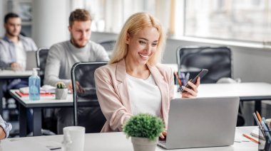 Menajer müşteriyle çevrimiçi ve sosyal mesafede çalışır. Gülümseyen kadın çalışan akıllı telefonu tutuyor ve modern ofis içindeki iş yerinde dizüstü bilgisayarda yazıyor. Karantinadan sonra geri dönüyor, boşluğu kopyalıyor.
