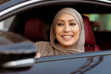 Mutlu gülümseyen Arap kadının yakın plan portresi, tesettürlü araba, sürücü koltuğunda oturan, yeni otomobilini kontrol eden, yan aynanın içinden yola bakan, dış görünüşü...
