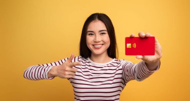 Neşeli genç bir kadın, bir barış işaretiyle parlak sarı bir arka plana karşı temassız bir ödeme kartını gururla sergilerken, alışverişte bir anlık mutluluk ve rahatlık sergiliyor..