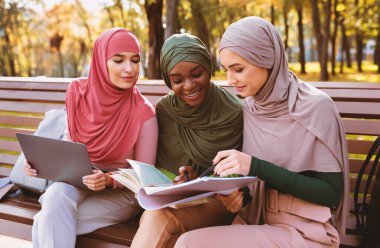 Üç Müslüman kadın öğrenci birlikte kitap okuyarak ve bilgisayar kullanarak dışarıda, kampüs parkında sıralarda oturuyor. Modern Arap Kadınları İçin Eğitim, Çevrimiçi Çalışma Konsepti