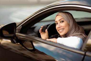 Kendi otomobili. Gülümseyen, başörtülü, araba anahtarlarını gösteren, yeni aracında oturan Müslüman bir kadının portresi. Kendinden emin Orta Doğulu Kadın Lüks Taşımacılığında Sürücü koltuğunda poz veriyor
