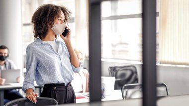 Müdür telefonda müşteriyle konuşuyor. Koruyucu maske takmış çekici Afro-Amerikan bayanı akıllı telefondan konuşurken aynı zamanda ofisin iç kısımlarındaki iş konularını tartışır.