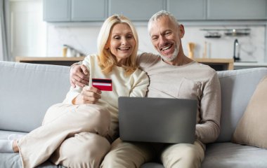 Neşeli yaşlı bir çift bir kanepeye yakın otururlar, online alışveriş yaparken gülümserler. Biri kredi kartı taşırken, diğeri dizüstü bilgisayar kullanarak evde samimi ve çekici bir ortam yaratıyor..