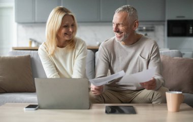 Gülümseyen yaşlı bir çift rahat bir oturma odasında dizüstü bilgisayarlarında işbirliği yapıyor, belgeleri birlikte gözden geçiriyorlar. Onların neşeli ifadeleri öğleden sonraki çalışma saatlerinde takım çalışmasını ve bağlılığını gösteriyor..