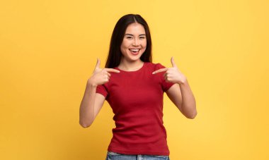 Sahnede genç bir Asyalı kadının sevinçle kendini göstermesi ve bir heyecan hissi vermesi yer alıyor. Kırmızı bir gömlek giyiyor. Parlak sarı arka planla çelişiyor..