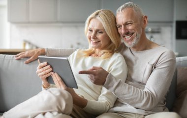 Mutlu bir son sınıf çifti rahat bir koltukta oturup tablete bakarken bir anı paylaşıyor. Açıkça nişanlanmışlar ve modern oturma odalarında birlikte vakit geçirmekten keyif alıyorlar..