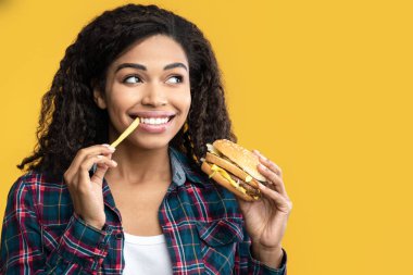 Fast Food. Gülümseyen Afro-Amerikan Kadın 'ın yakın plan portresi hamburger ve patates kızartması yiyen, fotokopi uzayına bakan. Zenci Bayan Tutma ve Isırma Lezzetli Patates, Portakal arka planda izole edilmiş, Banner