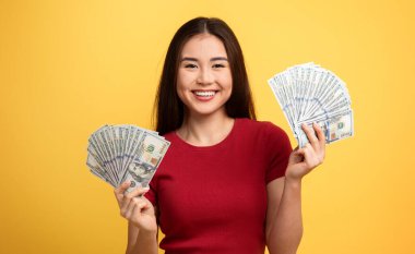 Genç bir Asyalı kadın gülümseyerek gururla iki elinde de yığınla para gösteriyor. Parayla ilgili heyecanını vurgulayarak neşeli tavırlarına hayat veren sarı arka plan ekleniyor..
