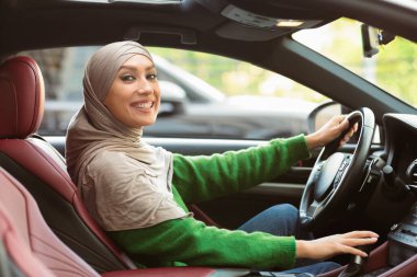 Araba sigortası konsepti. Gülümseyen Müslüman kadın sürücü modern araçta oturuyor, yeni arabasını sürmekten zevk alıyor, direksiyon ve vites kolu tutuyor, lüks salondaki kameraya poz veriyor.