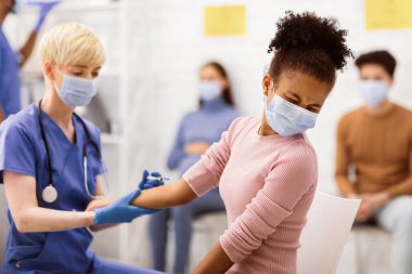 Coronavirus aşısı. Siyahi genç kız Covid-19 aşısı oluyor. Koluna enjekte ediliyor. Hastane odasında hemşireyle oturuyor. Corona Virüs Önleme, Tedavi ve İlaç Konsepti. Seçici Odaklanma