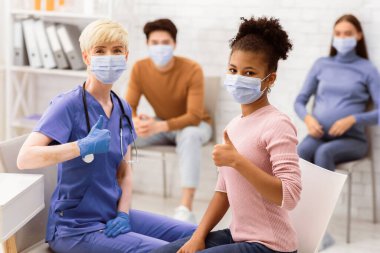 Başarılı Coronavirus tedavisi ve iyileşme sürecinden sonra hastane odasında maske takarak başparmak hareketi yapan kadın doktor ve Afrikalı hasta kız. Covid-19 İlaç Tedavisi ve Aşı