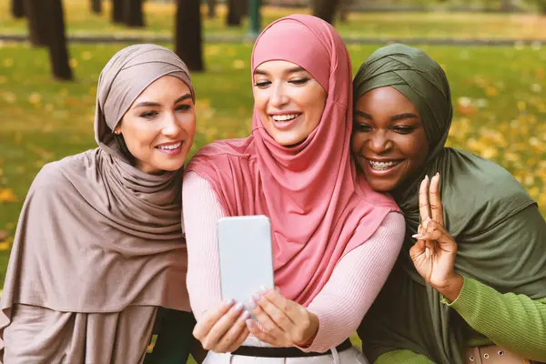 Üç modern Müslüman kadın telefonda bankta otururken eğlenerek selfie çekiyor. İslami Kadın Öğrenciler Hafta Sonu Dışarıda, Park 'ta Eğleniyor. Arkadaşlık Konsepti