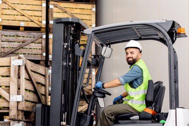 Elektrikli forkliftteki çalışan depoda olgun elmalarla dolu bir kap taşıyor. Gülümseyen milenyum sakallı adam kamyon kaskı takıyor, meyve kaldırıyor, depodaki ahşap sandıklar üzerinde, boş alan.