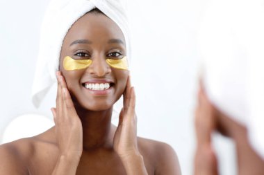 Sağlıklı, taze ciltli, yaşlanmayı önleyen göz maskesi, tedavi ve kozmetoloji. Göz altı kolajen altın pedleri olan mutlu Afro-Amerikan bayan, altın bantlarla aynaya bakıyor.