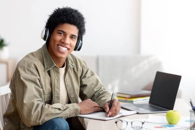 Web tabanlı eğitim. Kulaklıklı genç adam dizüstü bilgisayarda Webinar izlerken bilgi yazıyor, uzayı kopyalıyor. Havalı Afro-Amerikan gençliği çevrimiçi kursu için ev ödevi yapıyor