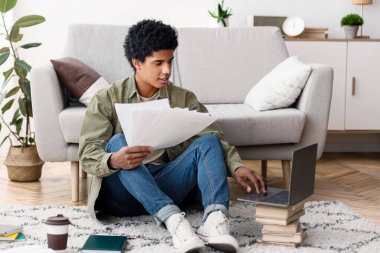 Young teen guy with educational materials, books and laptop computer working on cousework or school project at home. College student studying online, writing essay or graduation thesis
