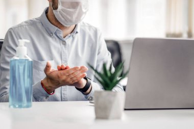 İş, yeni normal ve işyerinde sağlık koruması. Koruyucu maskeli genç adam elleri antiseptikle dezenfekte eder ve dizüstü bilgisayara bakar, tencerede bitkilerle birlikte boş bir masada oturur,