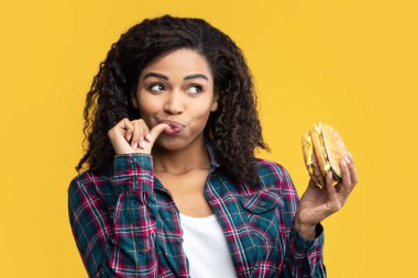 Hile Yemeği. Mutlu Afro-Amerikan kadın portresi Lezzetli Burger yiyor, parmağını yalıyor, fotokopi uzayına bakıyor. Fast food 'u elinde tutan tatmin olmuş kadın, turuncu arka planda izole edilmiş, afiş.