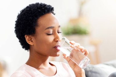 İyi bir sağlık konsepti için hidrasyon. Evdeki kanepede bir bardak berrak su içen güzel siyahi bir bayan. Afro-Amerikalı kadın köpüklü su içiyor, sağlığıyla ilgileniyor.