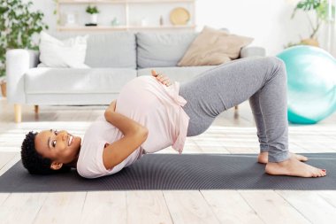 Evdeki yoga minderinde karın kası egzersizi yapan siyahi kadın köprü yogası pozunda duruyor, gelecekteki çocuğuna bakıyor. Genç hamile kadın sağlıklı bir hayat sürüyor, içeride çalışıyor.