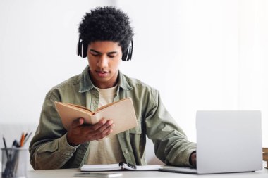 Evde dizüstü bilgisayar dersinde kulaklık takmış, kitap okuyan zenci bir öğrenci. Boş yer. Çevrimiçi öğrenim gören siyahi gençlik, üniversite hocasıyla webcam üzerinden iletişim kurmak.